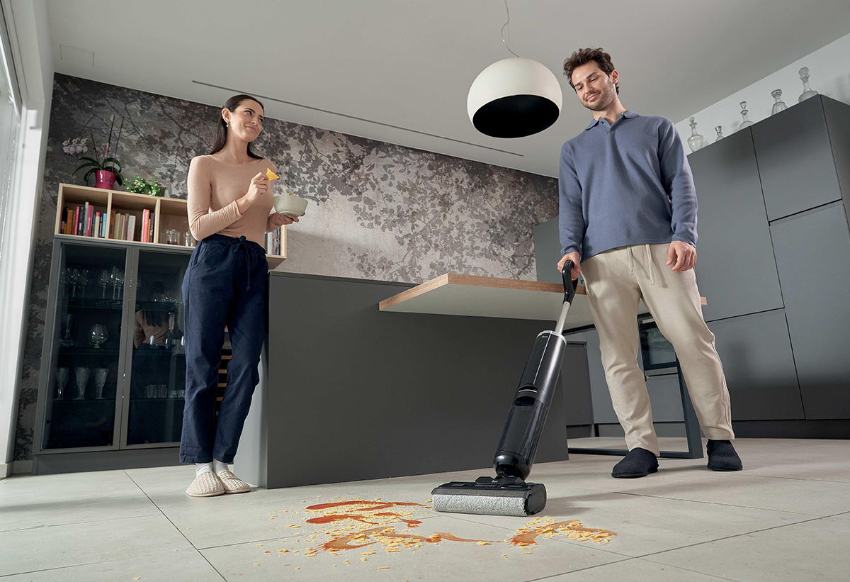 Απεικονίζεται ζευγάρι σε κουζίνα, ο άνδρας σκουπίζει.